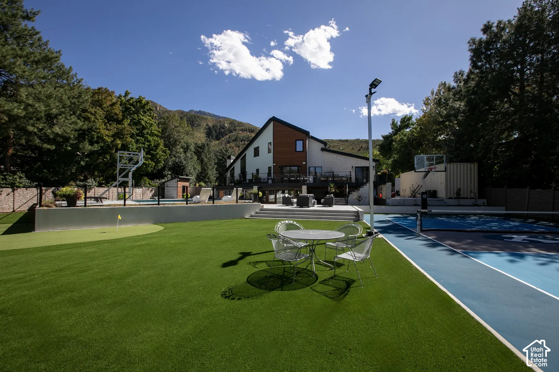 Exterior grounds featuring a synthetic turf putting green, a paved sports court, a swimming pool with glass panel fencing, a multi-level deck, and a modern residence with mixed-material siding