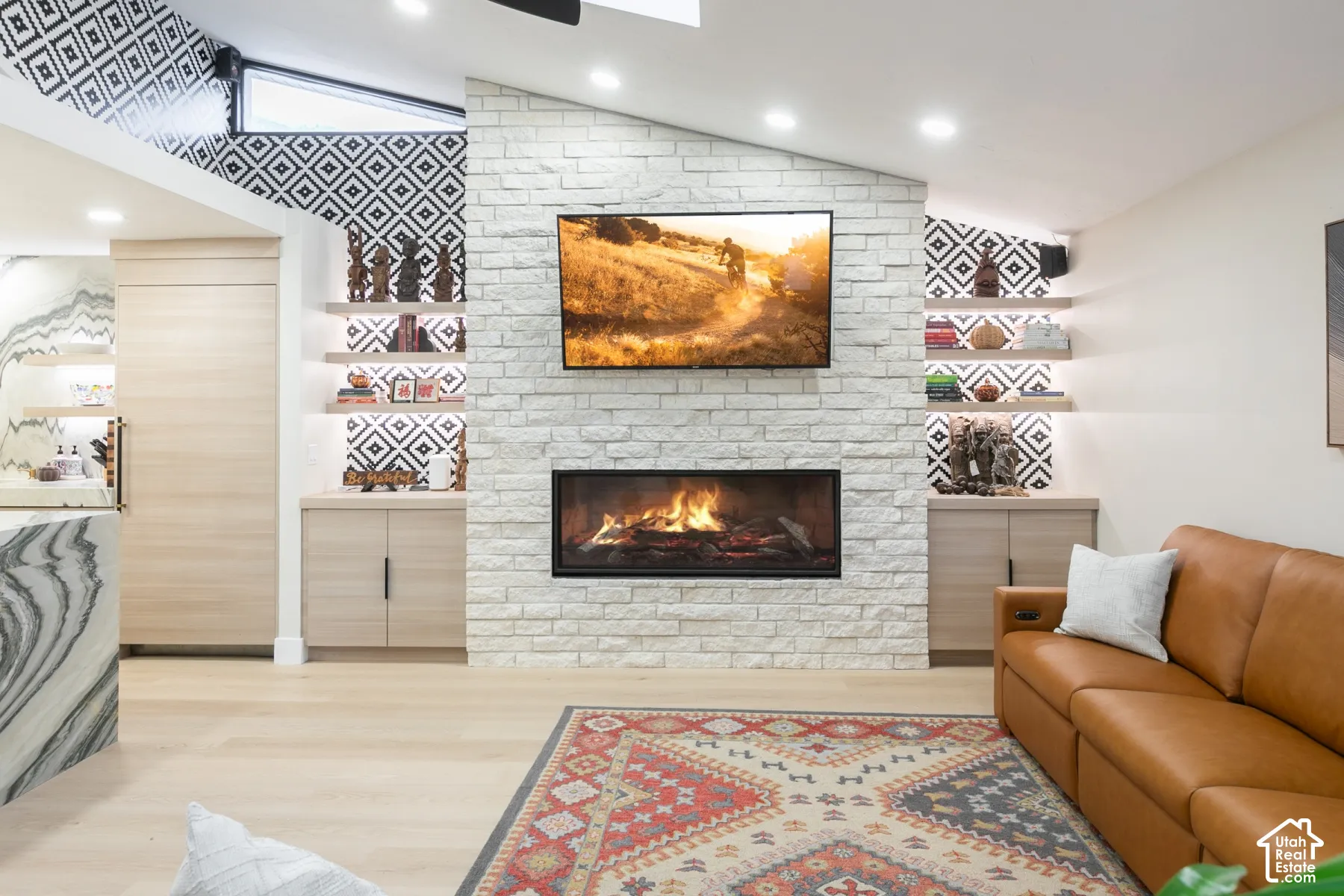 Stacked stone fireplace with a linear firebox, light wood-finish cabinetry, built-in shelving, patterned accent wall, and recessed ceiling lighting
