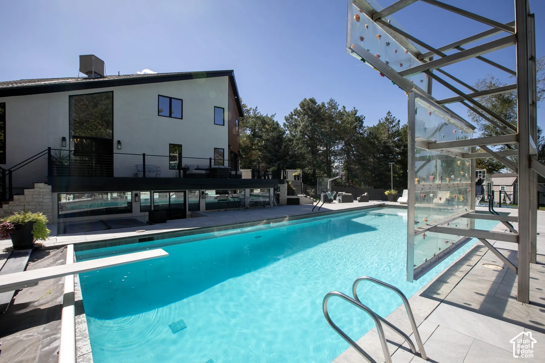 Swimming pool featuring a diving board, metal ladder, and built-in climbing wall, finished with light-toned stone decking