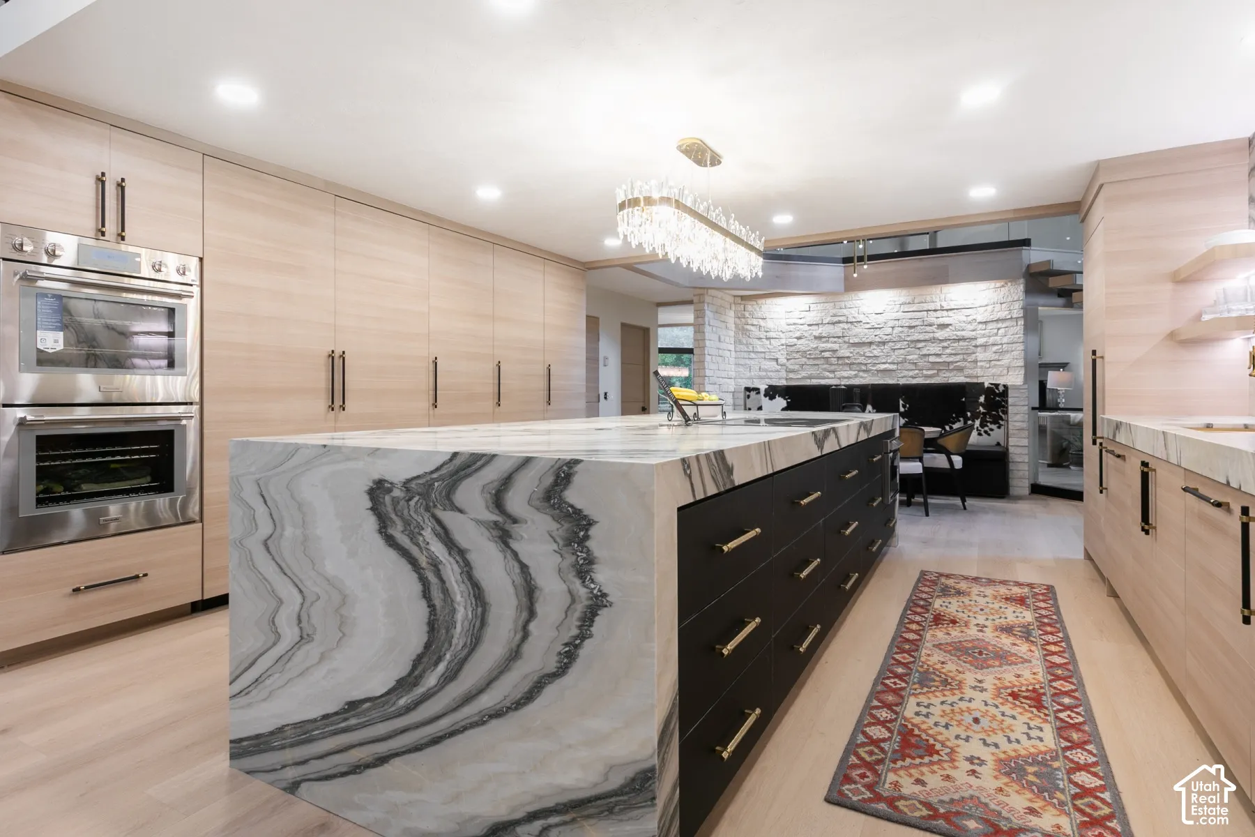 Kitchen featuring a waterfall-edge island with natural stone surfacing, matte black cabinetry with gold-tone hardware, wood-finish cabinetry, stainless steel double wall ovens, and a natural stone-clad accent wall