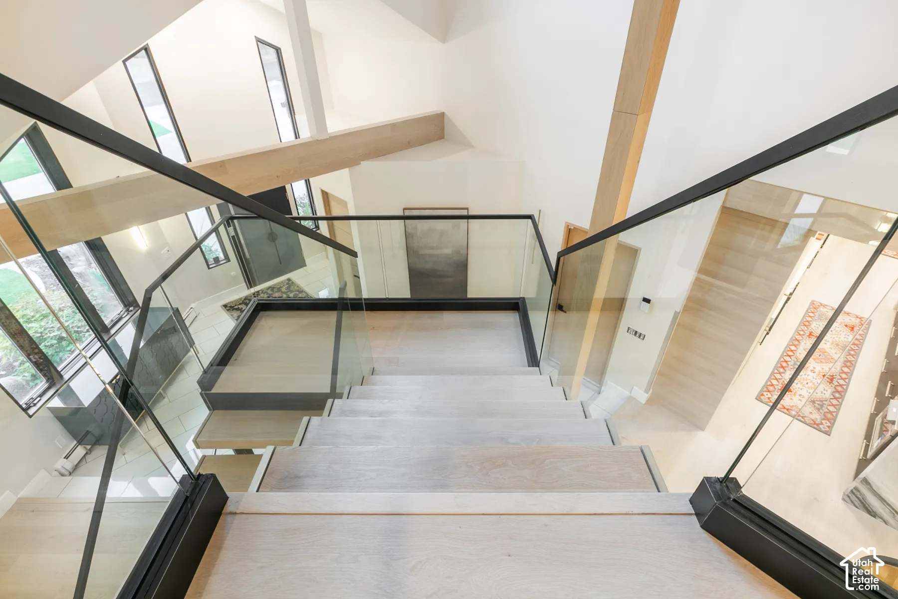 Staircase featuring wood-finish treads, glass panel railings, and matte black metal framing with exposed timber ceiling beams and vertical windows
