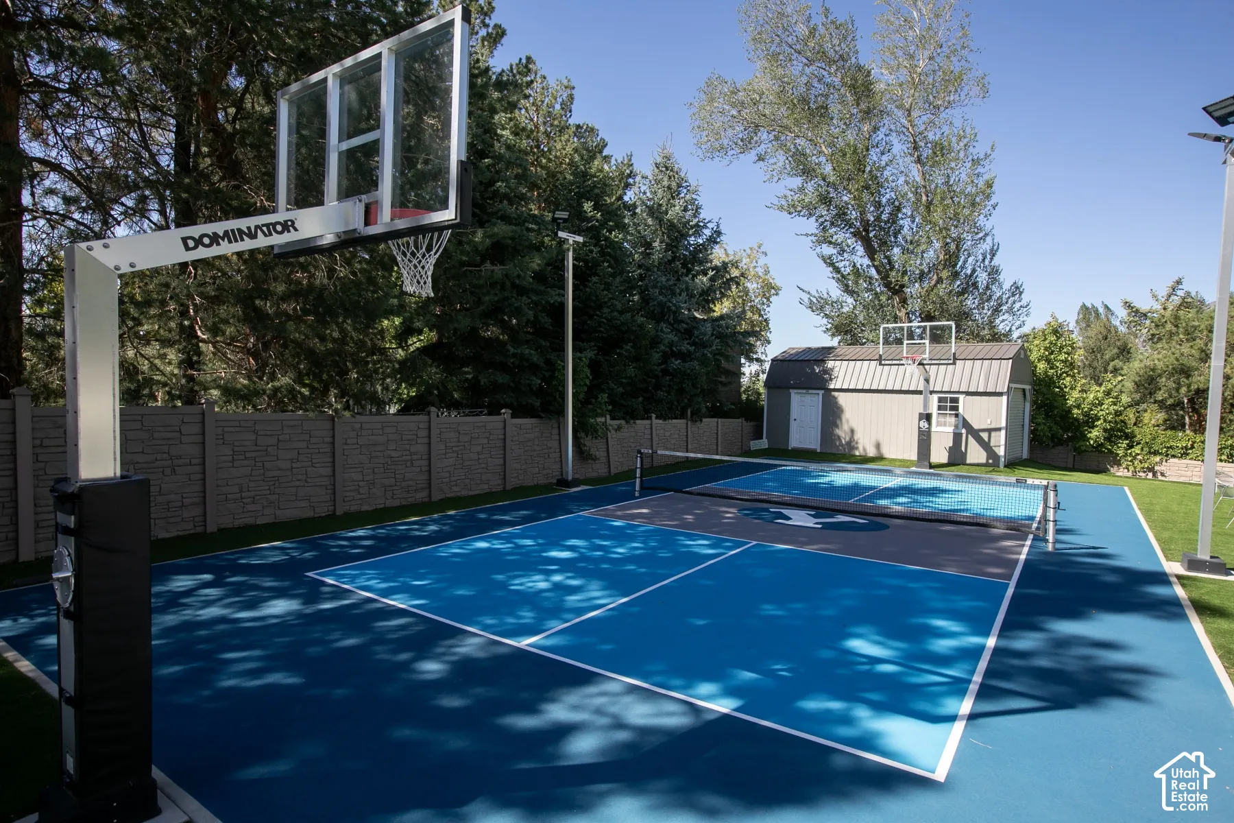 Multi-use sports court featuring a synthetic surface, basketball hoop system, net, stadium-style lighting, and enclosed by composite perimeter fencing with an external storage shed