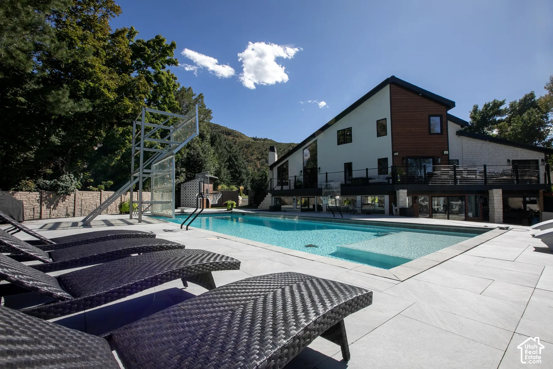 Swimming pool with a baja shelf, expansive stone-tile patio, integrated basketball hoop, elevated railed deck, and wood-accented siding