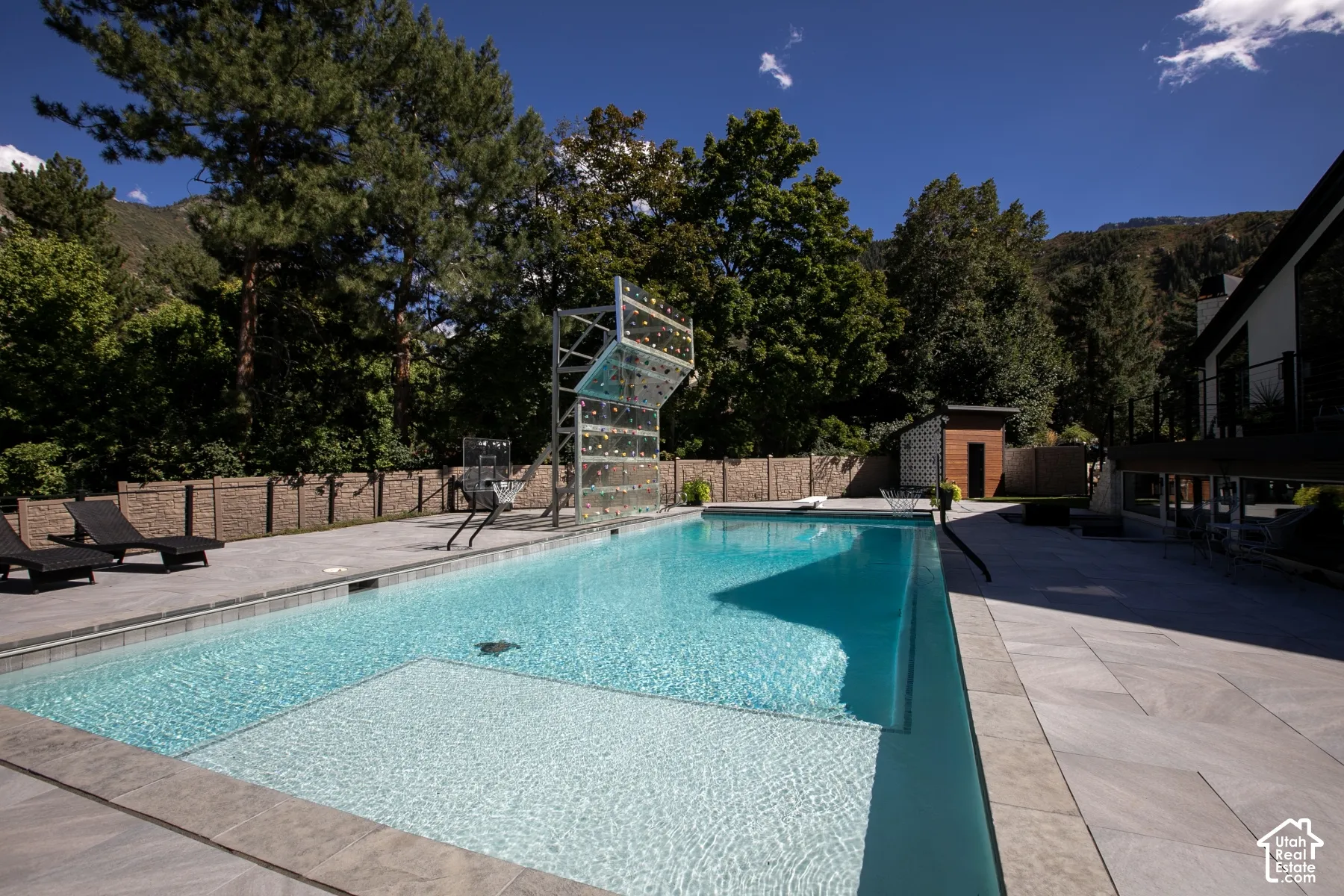 In-ground swimming pool with a Baja shelf, surrounding stone tile patio, built-in climbing wall structure, and perimeter privacy fencing