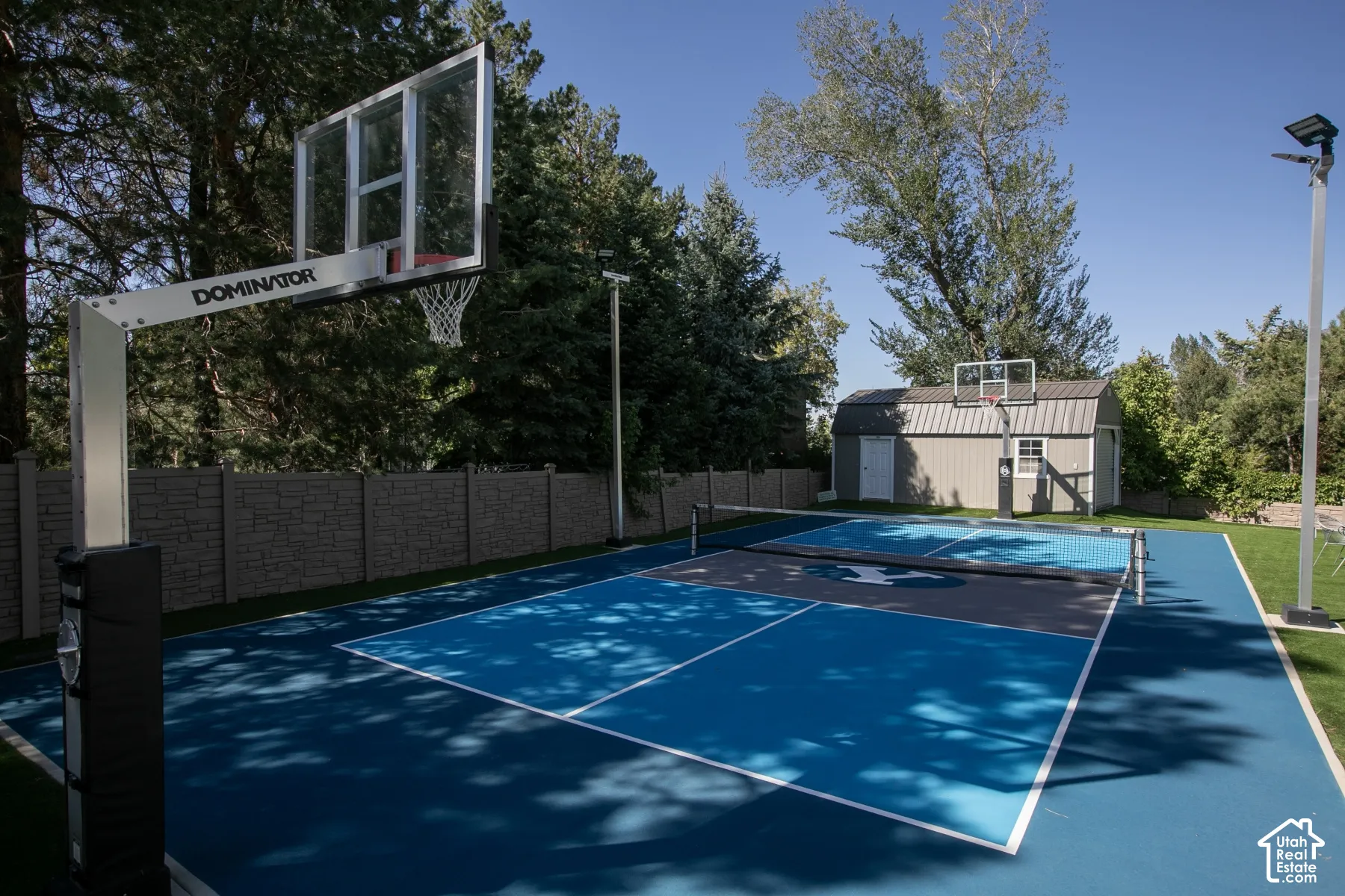 Multipurpose outdoor sports court featuring a dual-basketball system, net installation, specialized blue and grey court surface, perimeter fencing, and pole-mounted lighting