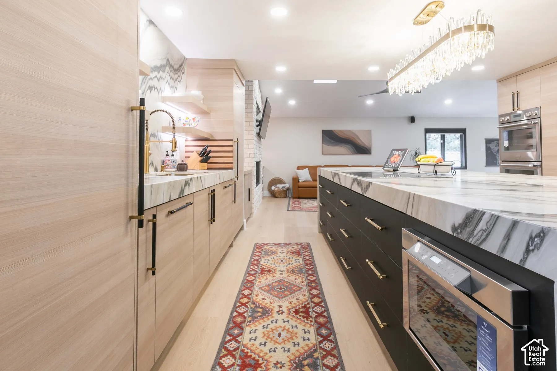 Kitchen featuring light wood-finish cabinetry, a large center island with stone-patterned surfacing, dark lower cabinetry, built-in stainless steel double ovens, and a statement chandelier