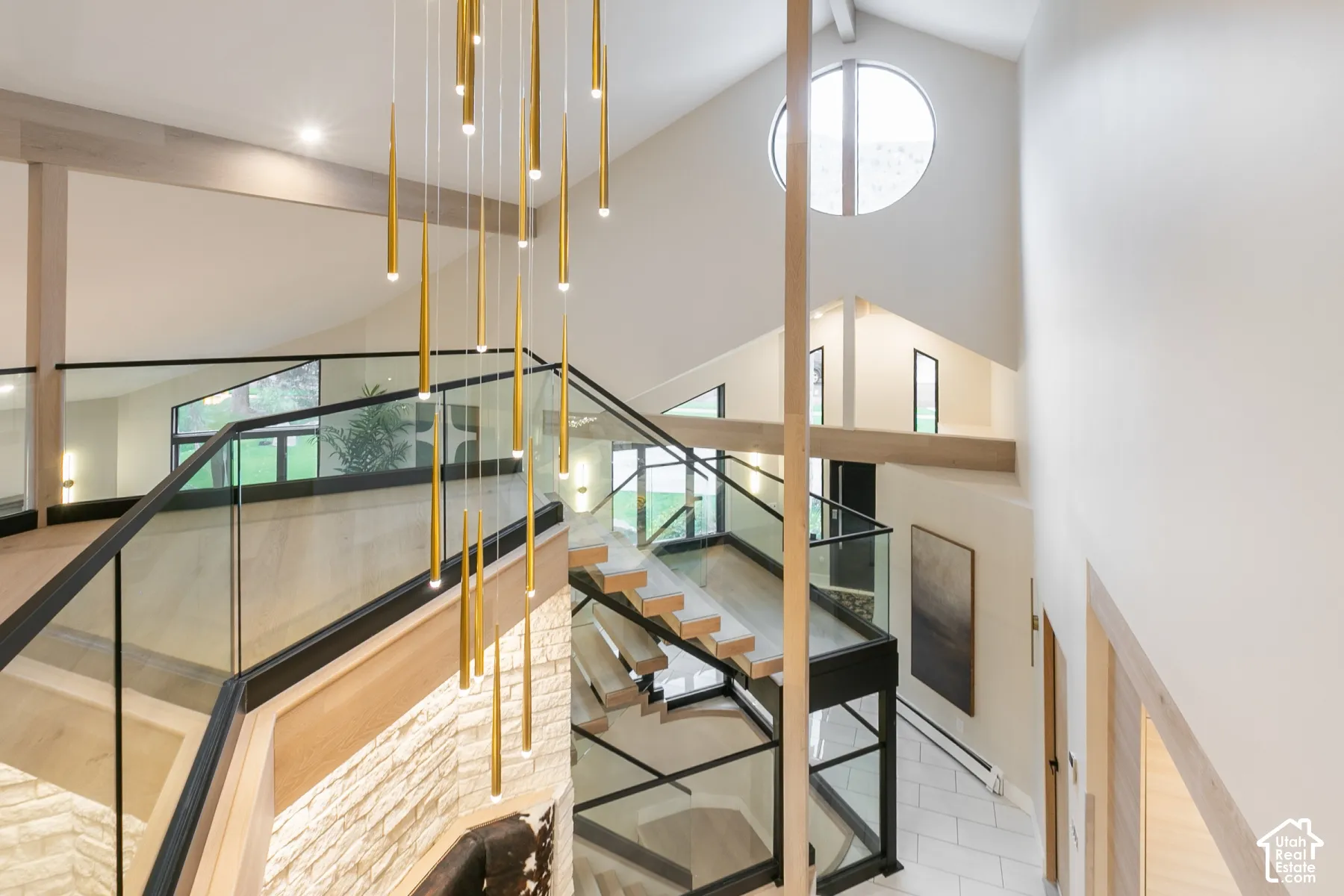 Multi-level staircase featuring open-tread wood stairs, glass railings with matte black framing, textured stone accent wall, circular clerestory window, and cascading gold-finish pendant lighting