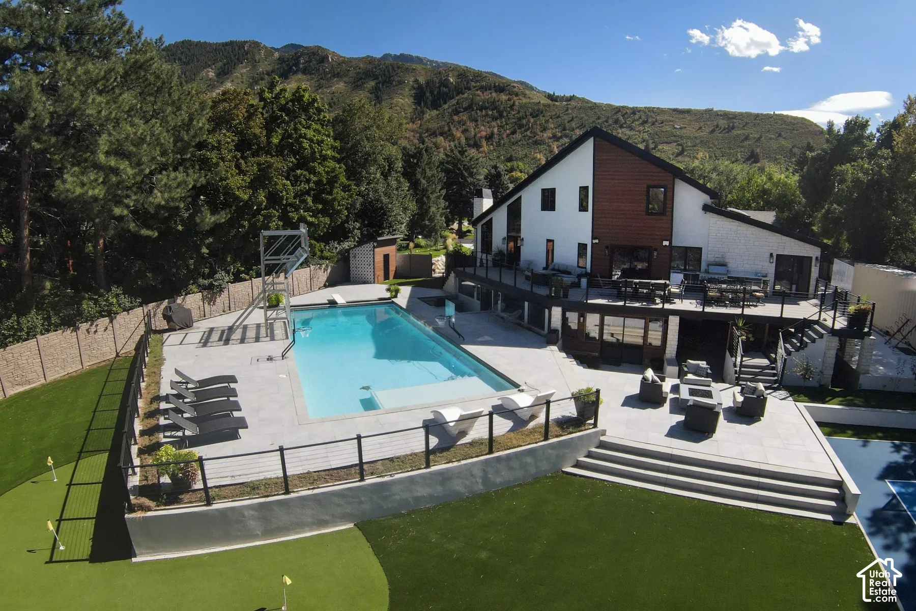 Multi-level exterior featuring wood-accented siding, an expansive stone-paved patio, an in-ground swimming pool with a diving board, and an integrated putting green, set against a mountainous backdrop