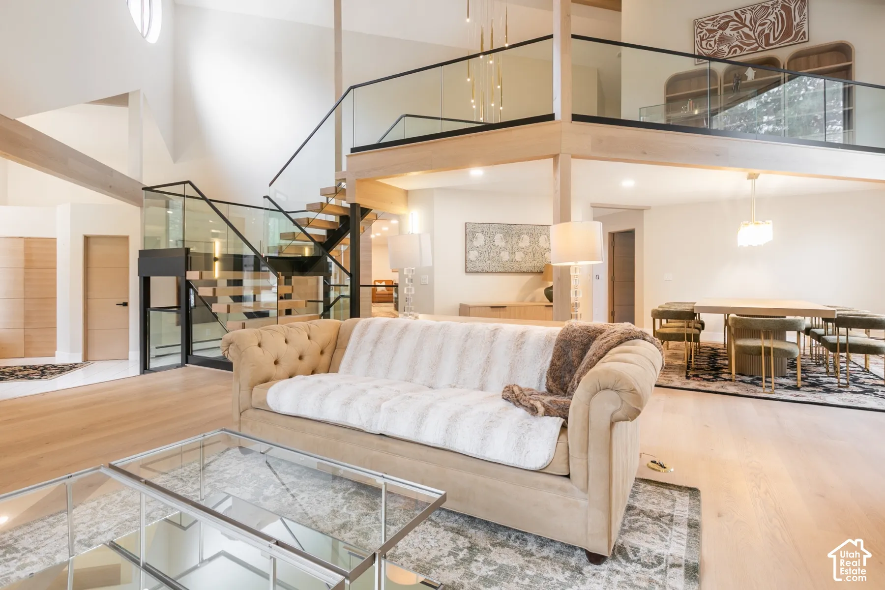Open-concept interior featuring light wood-finish flooring, vaulted ceilings, a structural staircase with glass railings, and a cantilevered mezzanine level