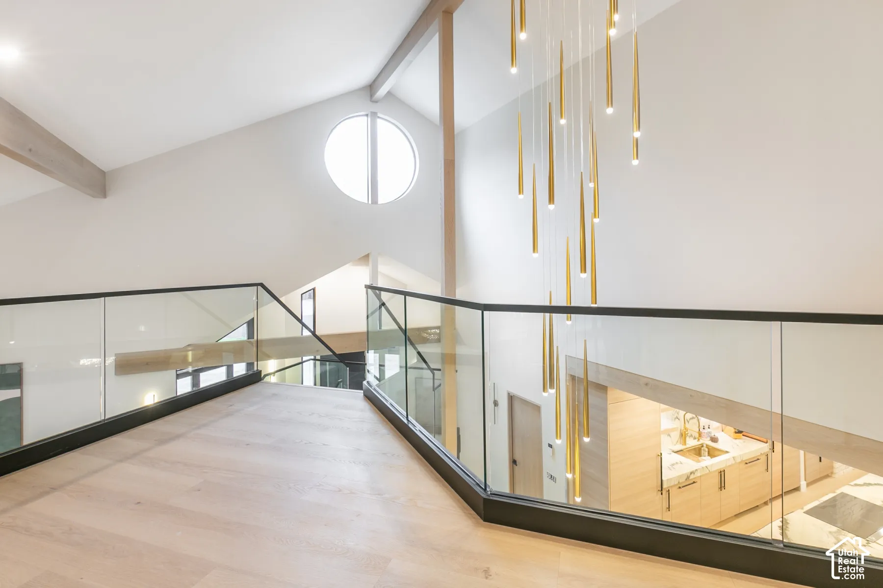 Vaulted ceiling featuring exposed wood beams, frameless glass railings with metal trim, circular window, and a multi-pendant brass light fixture