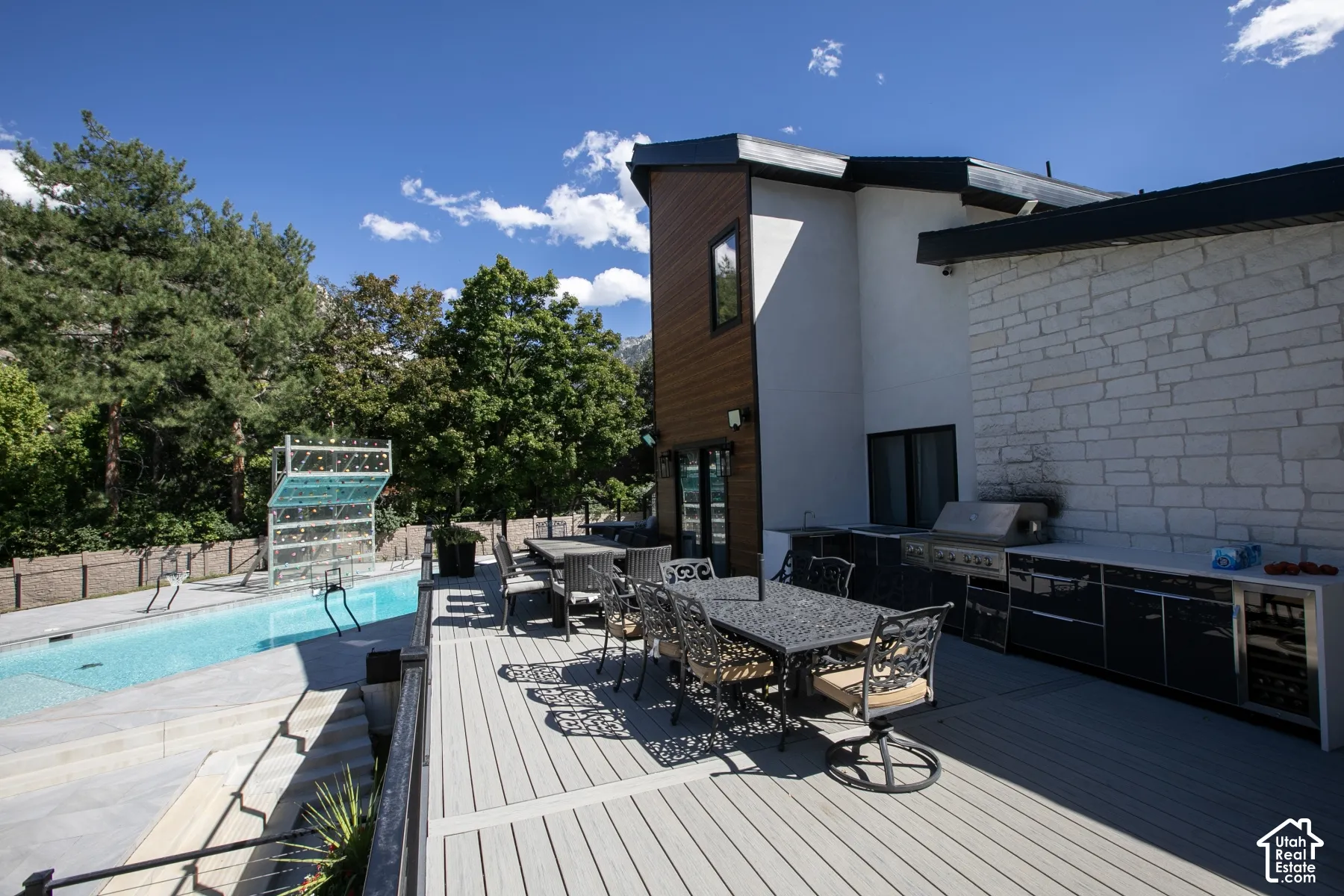 Multi-level exterior featuring wood-finish siding, white stone cladding, a synthetic deck, an in-ground swimming pool, and an outdoor kitchen with integrated stainless steel appliances