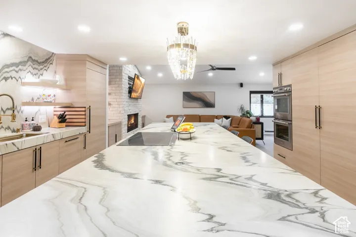 Kitchen featuring an oversized island with a stone countertop, flat-panel wood-finish cabinetry, built-in stainless steel double ovens, a decorative chandelier, and a floor-to-ceiling stone-tiled fireplace