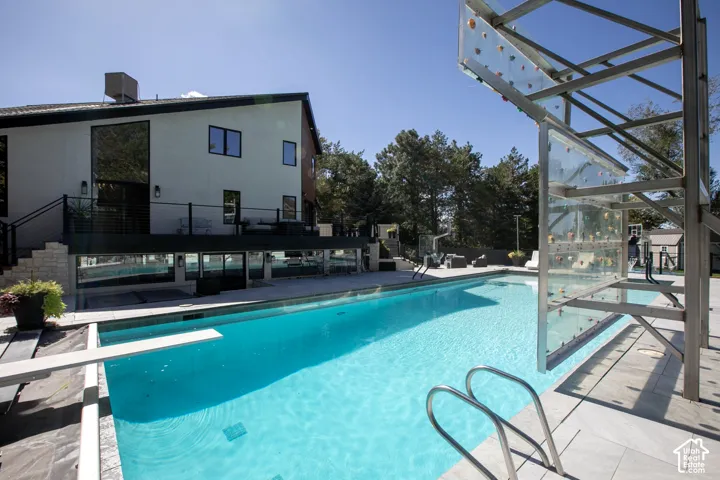 Swimming pool featuring a diving board, metal ladder, and built-in climbing wall, finished with light-toned stone decking