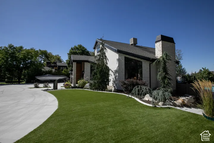Exterior featuring white stone siding, a prominent masonry chimney, high-contrast trim, expansive paved motor court, and landscaped lawn with ornamental trees