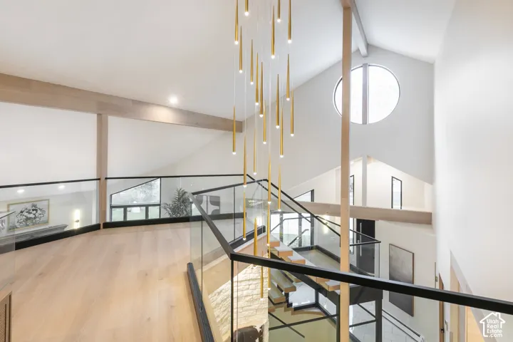Vaulted ceiling with exposed wood-finish beams, glass panel railings, tiered brass chandelier, circular window, and wood-finish flooring