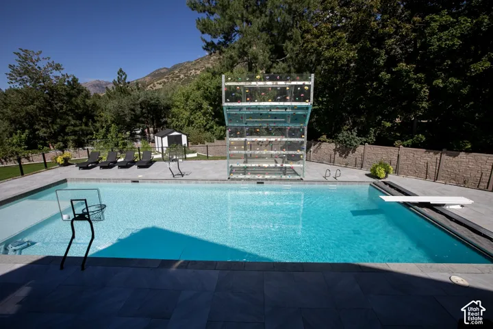In-ground swimming pool with a diving board, integrated climbing wall, expansive stone-paver decking, and perimeter stone-textured privacy fencing