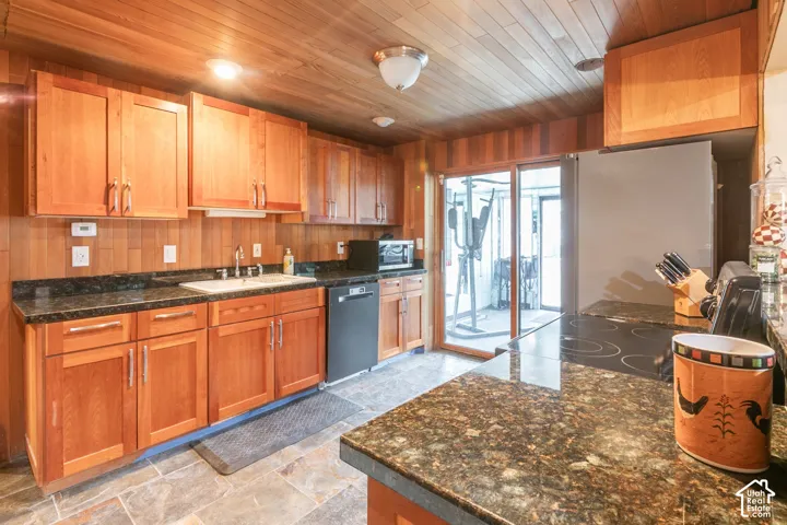 Kitchen featuring wood cabinetry, natural wood-paneled walls and ceiling, stone-finish flooring, and dark slab countertops with an integrated sink