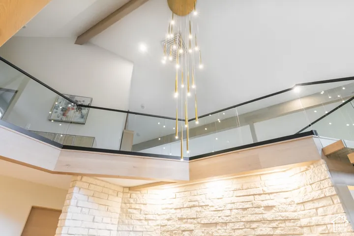 Vaulted ceilings featuring exposed wood beams, a stacked stone accent wall, a multi-pendant light fixture, and a glass-panel railing with a matte black finish