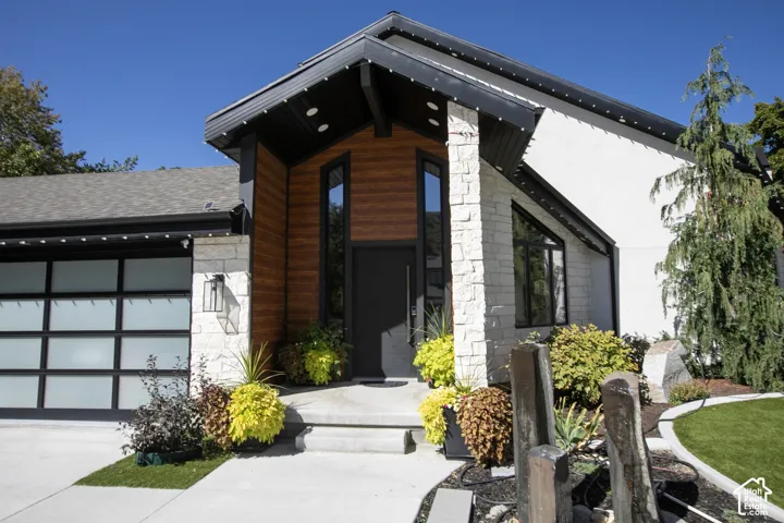 Modern exterior featuring stacked stone masonry, horizontal wood-finish siding, a frosted glass panel garage door, steep gable roofline, and vertical accent windows