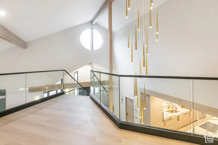 Vaulted ceiling featuring exposed wood beams, frameless glass railings with metal trim, circular window, and a multi-pendant brass light fixture