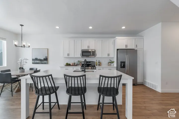 Kitchen featuring white shaker-style cabinetry, quartz countertops, central island with integrated sink, stainless steel appliances, and wood-finish flooring