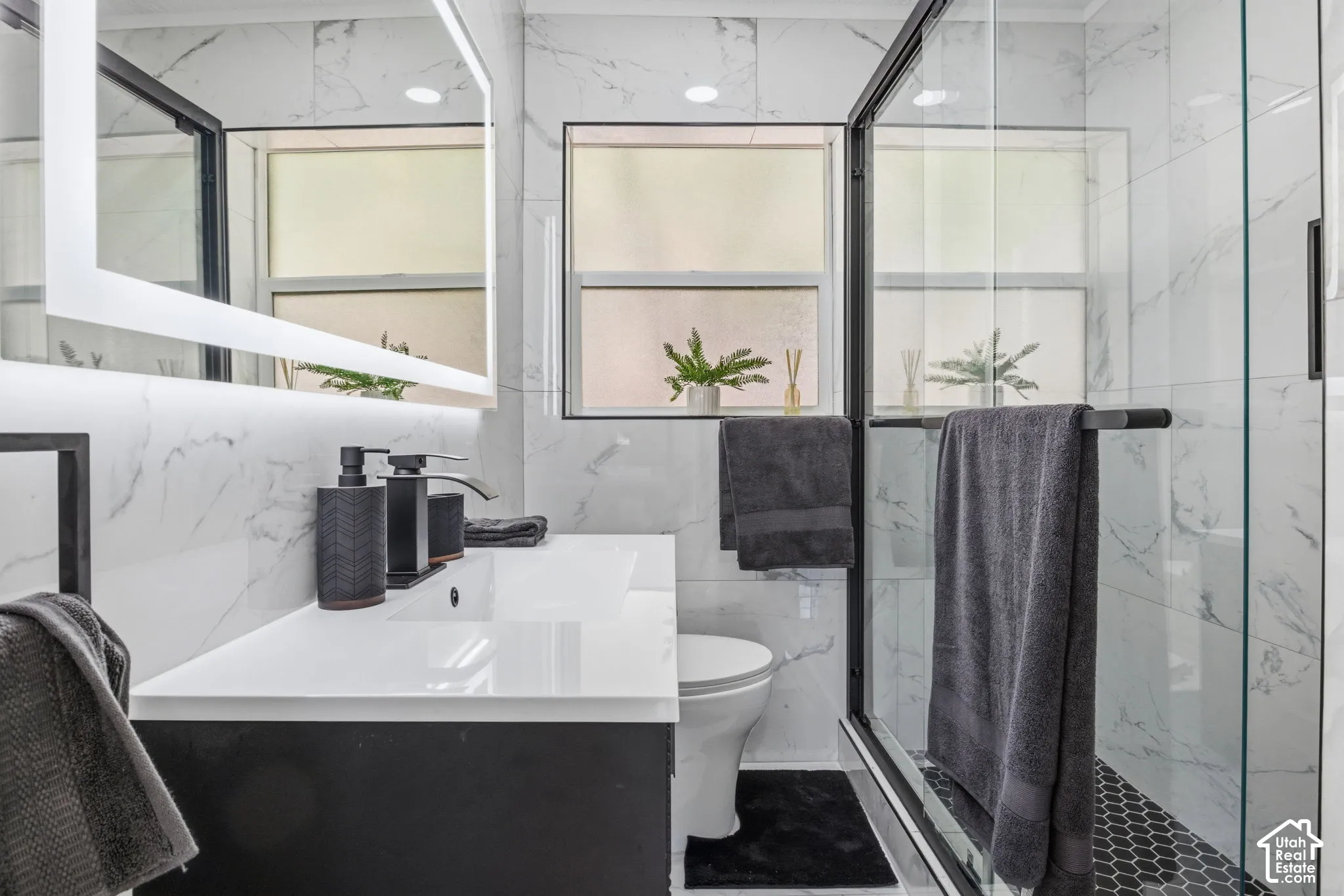 Modern vanity with a sleek white countertop, wall-to-wall LED-backlit mirror, floor-to-ceiling veined tile wall finishes, glass-enclosed shower with black metal framing, and geometric black floor tiling