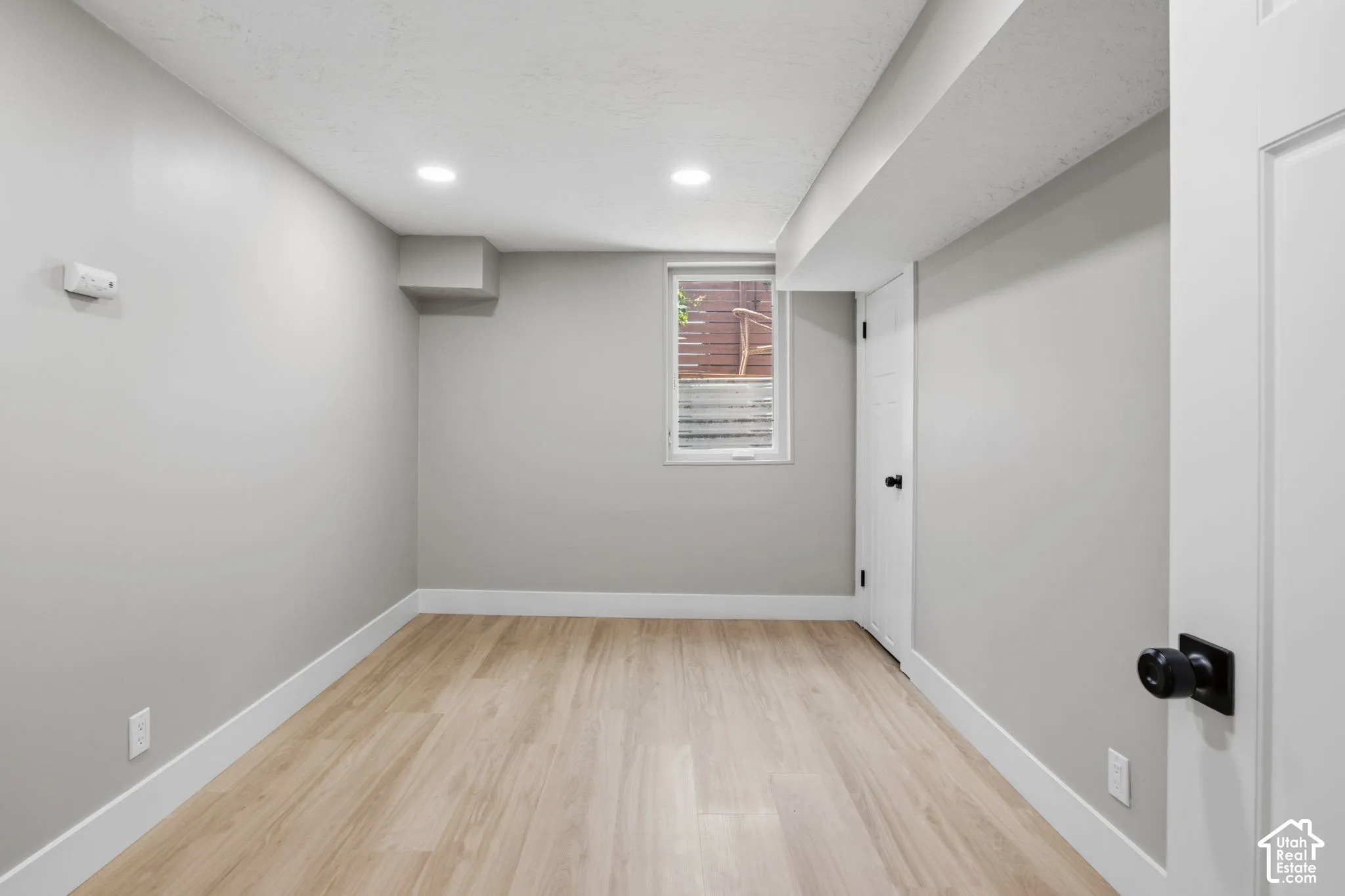 Neutral-toned interior space featuring wood-finish flooring, recessed lighting, a single pane window, and a white interior door with matte black hardware
