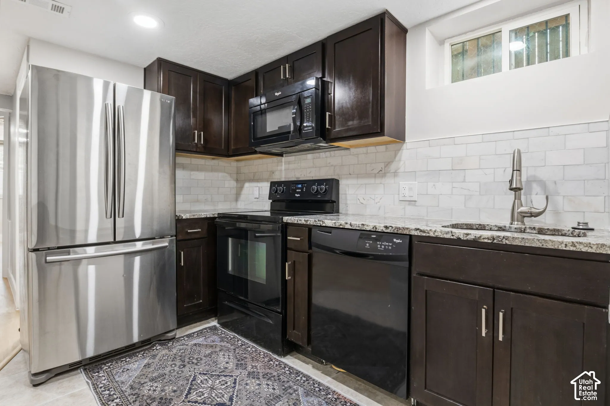 Kitchen featuring dark wood cabinetry, stone-finish countertops, subway tile backsplash, stainless steel refrigerator, and black built-in appliances