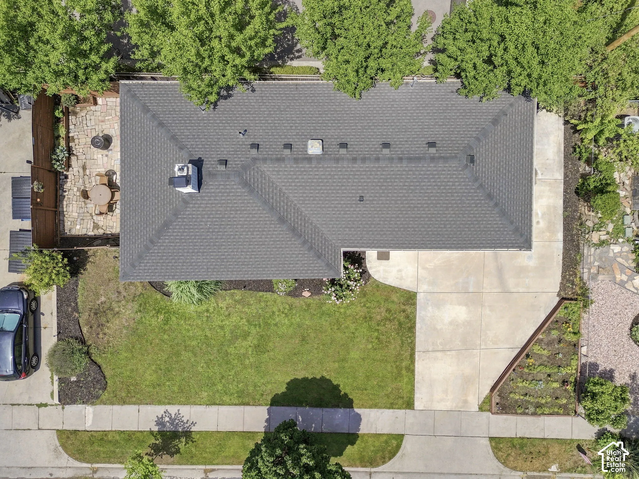 Aerial perspective featuring a shingled roof, concrete driveway, stone-paved patio, manicured lawn, and landscaped garden beds