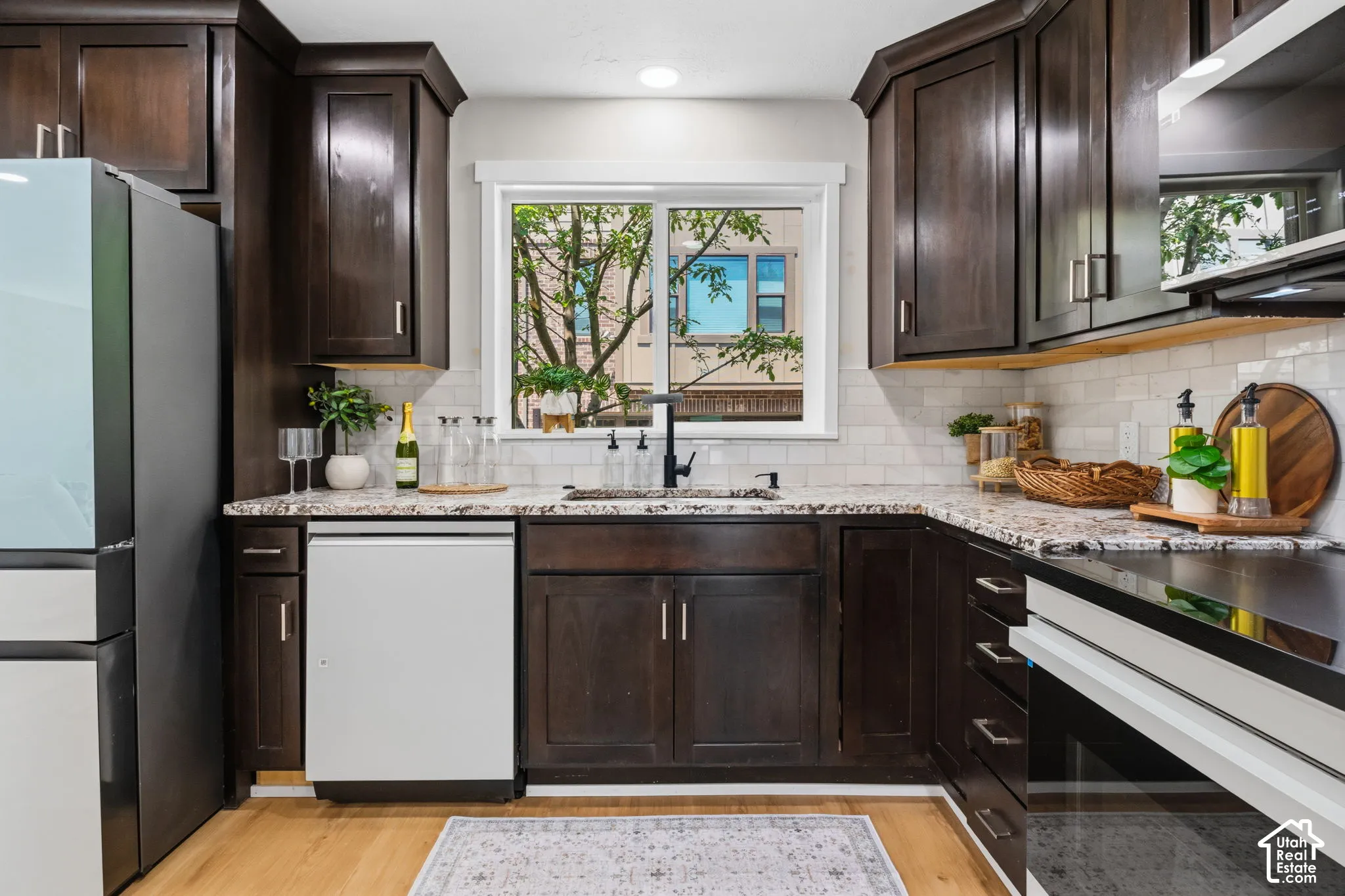 Kitchen featuring dark-finish cabinetry, patterned stone countertops, a white subway tile backsplash, a matte black kitchen faucet, and wood-finish flooring
