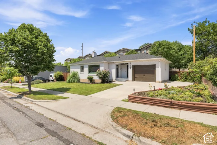 Single-story exterior featuring a painted brick facade, attached one-car garage, concrete driveway, and tiered garden beds