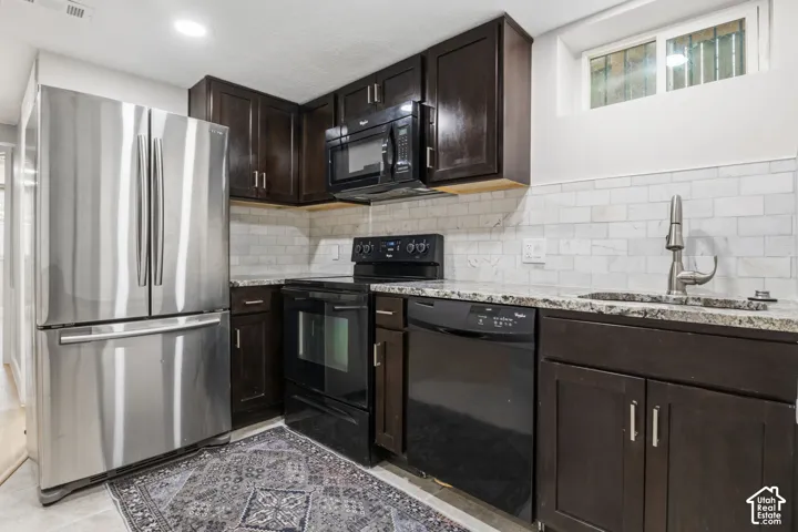 Kitchen featuring dark wood cabinetry, stone-finish countertops, subway tile backsplash, stainless steel refrigerator, and black built-in appliances