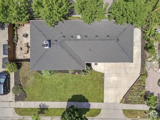 Aerial perspective featuring a shingled roof, concrete driveway, stone-paved patio, manicured lawn, and landscaped garden beds