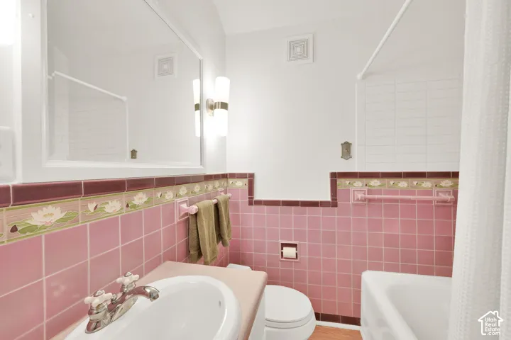 Bathroom featuring mid-tone pink wall tiling, decorative floral tile border, white drop-in sink with dual-handle metal faucet, wall-mounted light fixture, and large rectangular vanity mirror