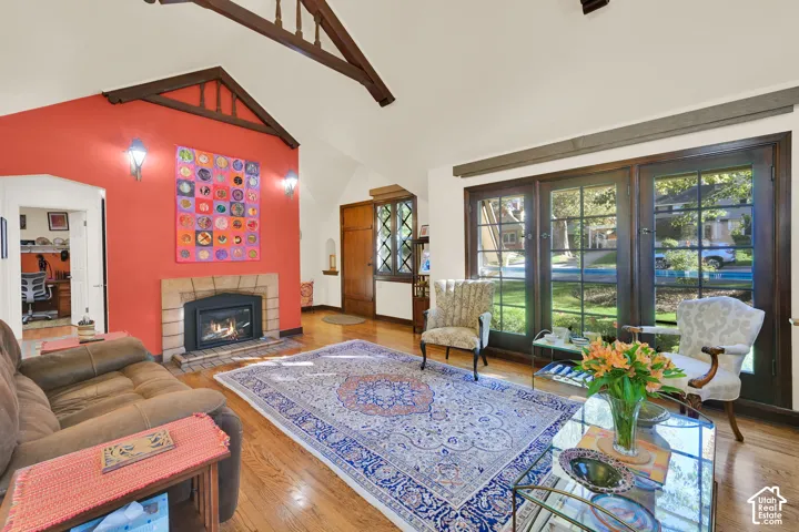 Vaulted ceiling with exposed wooden trusses, fireplace with stone surround, hardwood flooring, and multi-light glass paneled doors
