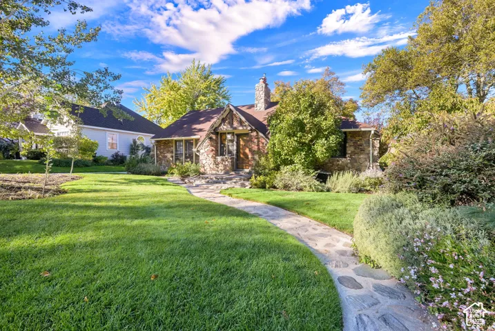 Stone exterior facade featuring a prominent stone chimney, arched entryway, and a winding flagstone walkway surrounded by manicured lawn and mature landscaping