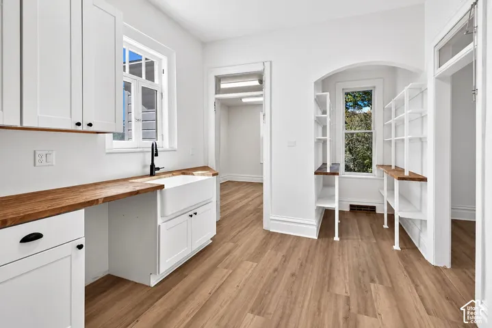 White cabinetry featuring wood-finish countertops, apron-front farmhouse sink with matte black fixture, and built-in shelving within an arched alcove, all set upon wood-finish flooring
