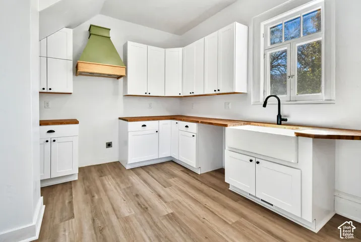 Kitchen featuring white shaker-style cabinetry, wood-finish countertops, apron-front sink with matte black faucet, custom range hood, and wood-finish flooring