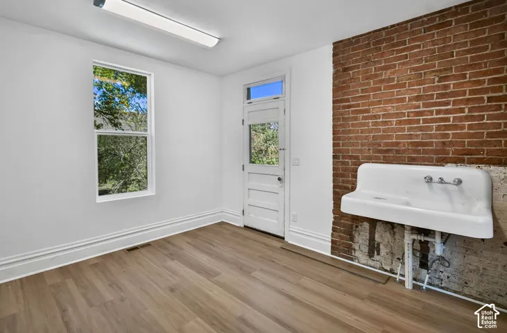 Exposed brick wall featuring a wall-mounted utility sink with chrome fixtures, wood-finish flooring, and a glass-paneled exterior door with transom window