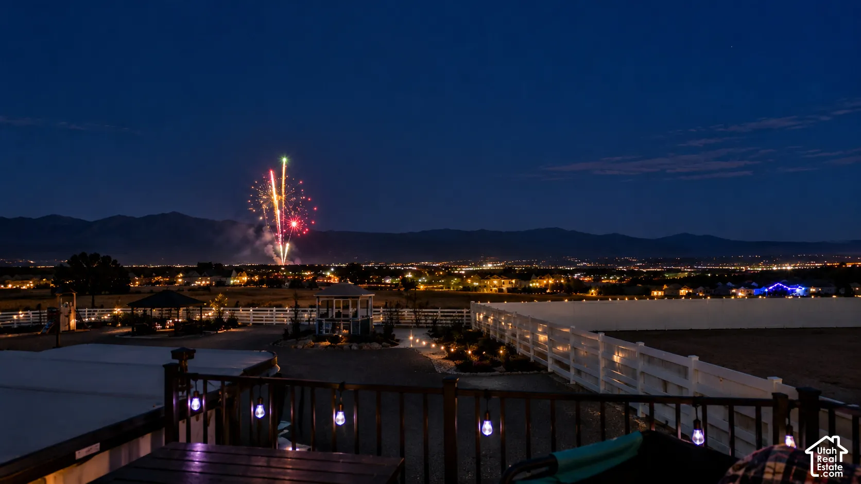 Outdoor deck featuring a wood railing with integrated string lighting, overlooking a white-fenced yard with a gazebo, set against a backdrop of mountain silhouettes and expansive horizon scenery