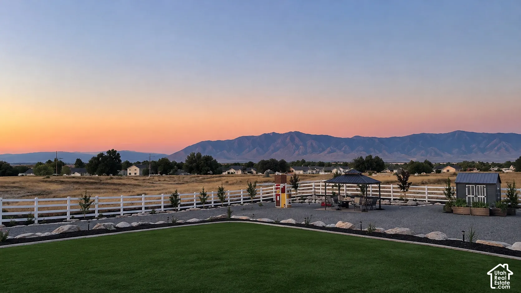 Fenced backyard featuring a manicured lawn, stone hardscaping, a metal-roof gazebo, and a storage shed, set against a mountain backdrop