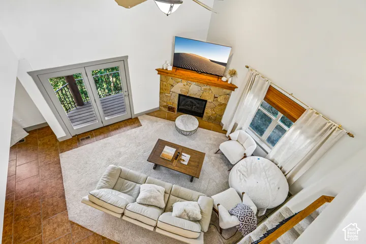 Double-height living area featuring a floor-to-ceiling stone fireplace with a wood mantel, tile flooring, and glass-paned double doors leading to an outdoor deck