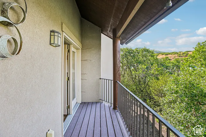 Covered balcony featuring wood-finish decking, dark metal railings, an overhanging wood-beam ceiling, wall-mounted exterior lighting, and a dual-door entrance