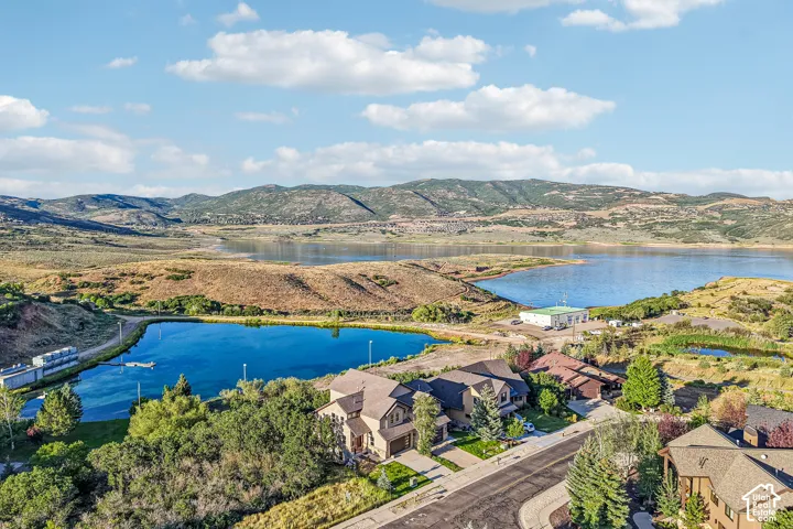 Aerial perspective featuring a multi-story residence with a multi-car garage, shingled roof, mountain backdrop, and proximity to a large body of water