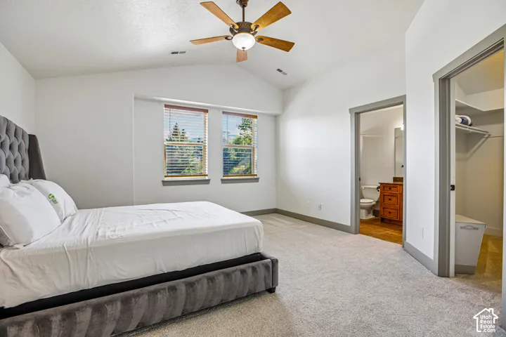 Room featuring vaulted ceilings, neutral carpeting, dual windows with blinds, a ceiling fan, and access to a bathroom and walk-in closet