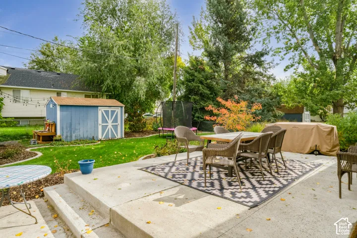 Backyard featuring a concrete patio, lawn area, and a detached storage shed with a shingle roof, surrounded by mature trees