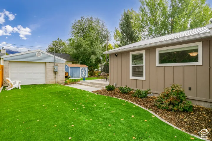 Rear exterior featuring board-and-batten siding, a metal roof, a detached garage with a paneled door, a concrete patio, and synthetic turf
