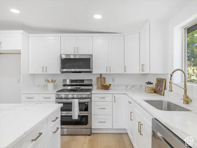 Kitchen featuring white shaker-style cabinetry, quartz countertops with integrated backsplash, stainless steel appliances, a brass gooseneck faucet, and light-tone wood-finish flooring