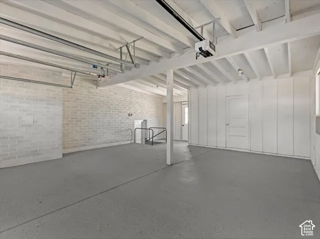 Attached garage featuring finished painted brick and paneled walls, painted concrete flooring, exposed beam ceiling, and an automatic garage door opener