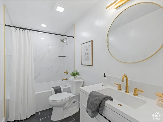 Bathroom featuring a white vanity with an integrated basin, gold-finish faucet, circular gold-rimmed mirror, and tub-shower surround with light-toned surfacing and gold hardware